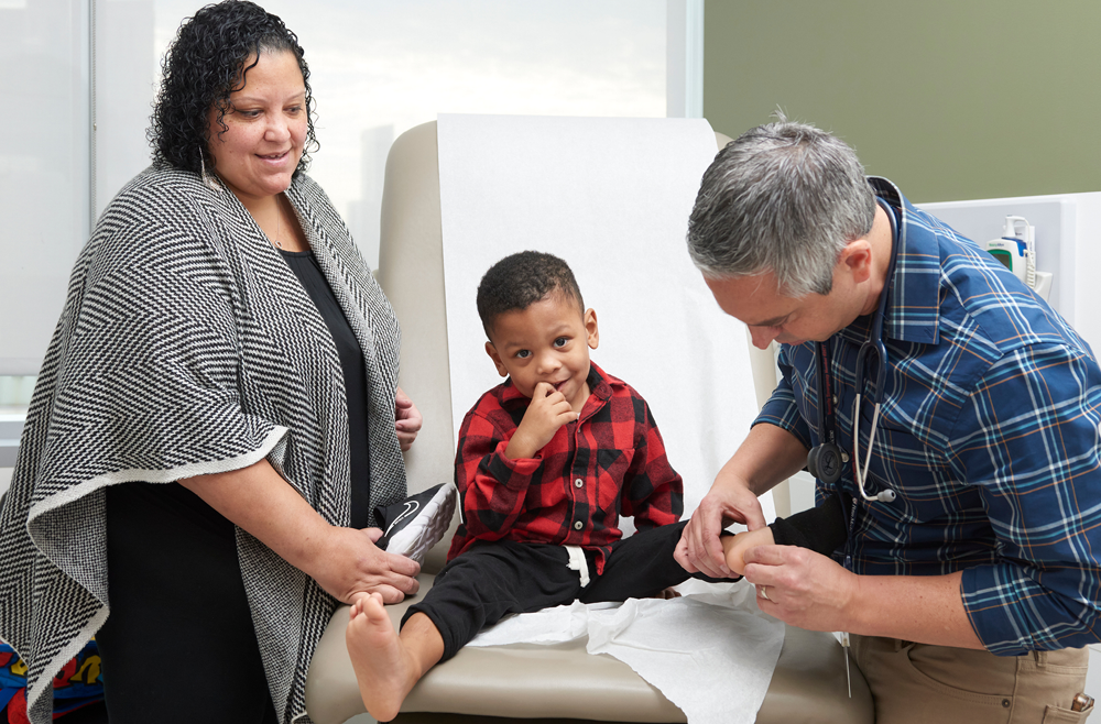 Nicole Miller and 3-year-old Elijah Miller in an exam room with Elijah’s doctor Mario DeMarco, MD checking on the child’s feet as he smiles.